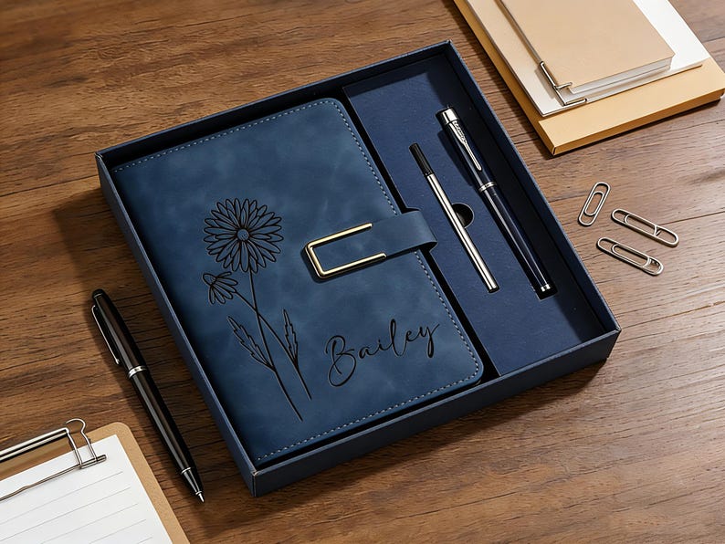 Puede incluir: Un juego de diario de cuero azul con un dise&ntilde;o floral y el nombre "Bailey" grabado en la portada. El juego incluye dos bol&iacute;grafos, un portal&aacute;pices y clips, todo presentado en una caja azul oscuro.