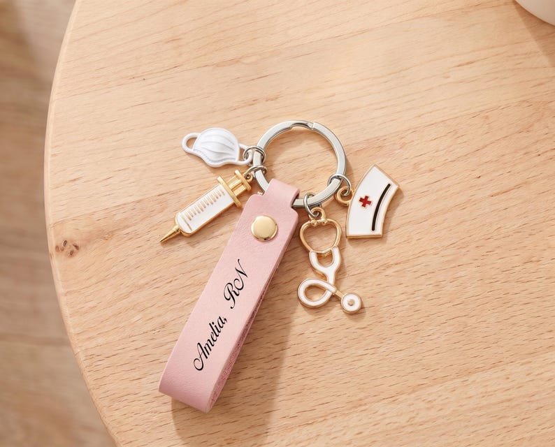 Pu&ograve; includere: Un portachiavi in pelle rosa con il nome "Amelia RN" inciso. Il portachiavi presenta ciondoli a forma di siringa, un cappello da infermiera con una croce rossa, uno stetoscopio e una mascherina chirurgica, tutti attaccati a un portachiavi argentato.