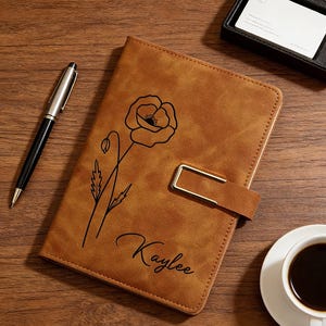 Puede incluir: Un diario de cuero marr&oacute;n con un dise&ntilde;o de flor de amapola negra y el nombre "Kaylee" se muestra sobre una superficie de madera. Tambi&eacute;n hay un bol&iacute;grafo negro y plateado, un tarjetero negro y una taza de caf&eacute;.