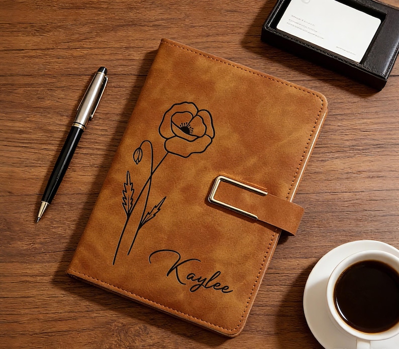 Puede incluir: Un diario de cuero marr&oacute;n con un dise&ntilde;o de flor de amapola negra y el nombre "Kaylee" se muestra sobre una superficie de madera. Tambi&eacute;n hay un bol&iacute;grafo negro y plateado, un tarjetero negro y una taza de caf&eacute;.