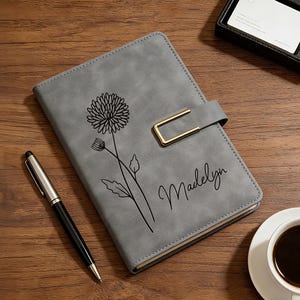 Puede incluir: Un diario gris con un dise&ntilde;o floral y el nombre "Madelyn" en cursiva. El diario tiene un cierre dorado y un bol&iacute;grafo est&aacute; al lado. Tambi&eacute;n se ven una taza de caf&eacute; y una peque&ntilde;a caja negra.
