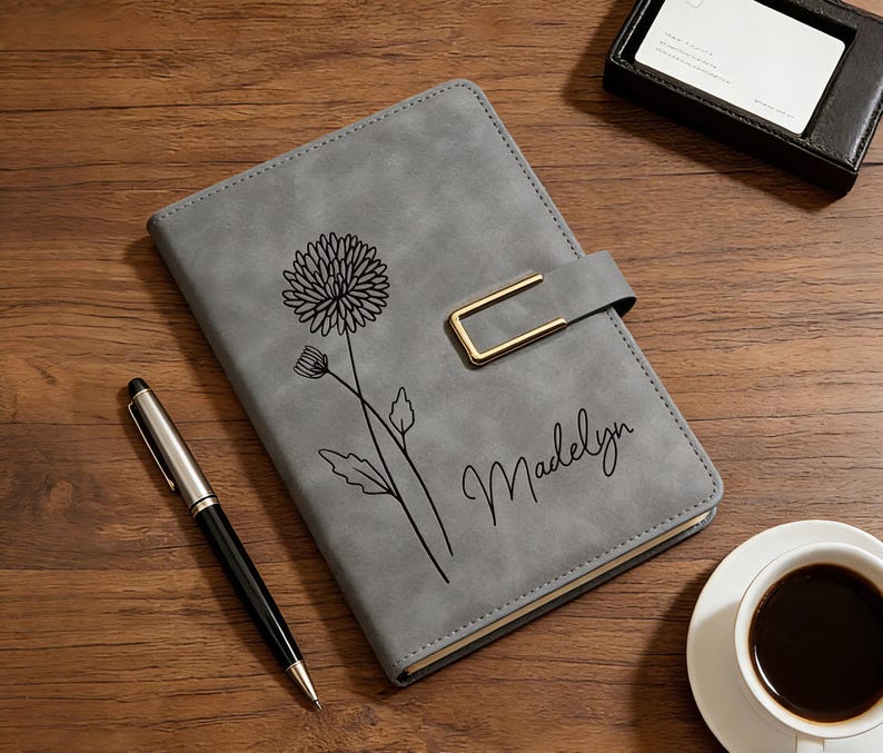 Puede incluir: Un diario gris con un dise&ntilde;o floral y el nombre "Madelyn" en cursiva. El diario tiene un cierre dorado y un bol&iacute;grafo est&aacute; al lado. Tambi&eacute;n se ven una taza de caf&eacute; y una peque&ntilde;a caja negra.