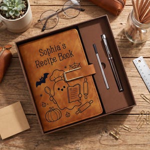 Puede incluir: Un libro de recetas de cuero sintético color canela con un cierre de metal dorado, con el texto "Sophia's Recipe Book" y dibujos lineales negros de un fantasma, murciélago, calabaza y utensilios de cocina. Incluye un bolígrafo plateado en la caja.