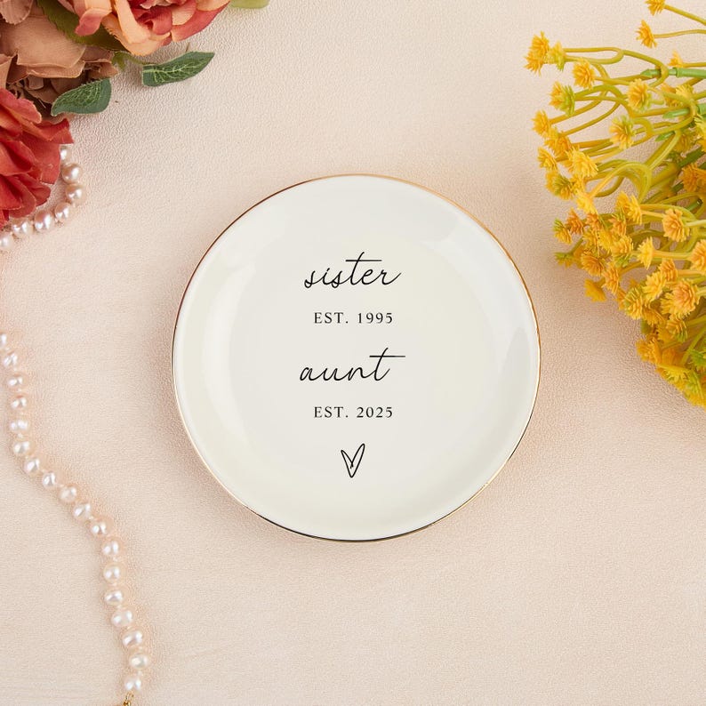 May include: A cream-colored ceramic trinket dish with a gold rim. The dish has the words "sister EST. 1995" and "aunt EST. 2025" in black script, with a heart symbol below. Decorative flowers and a pearl necklace are in the background.