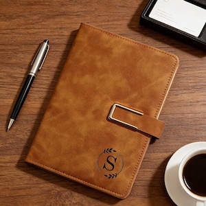 Op de afbeelding: Een bruin leren notitieboek met een gouden sluiting en de letter "S" in een lauwerkrans. Een zwarte en zilveren pen, een visitekaarthouder en een kopje koffie liggen op een houten oppervlak.