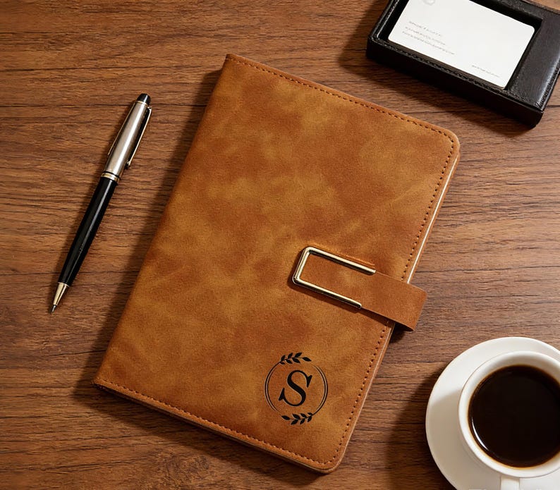 Op de afbeelding: Een bruin leren notitieboek met een gouden sluiting en de letter "S" in een lauwerkrans. Een zwarte en zilveren pen, een visitekaarthouder en een kopje koffie liggen op een houten oppervlak.