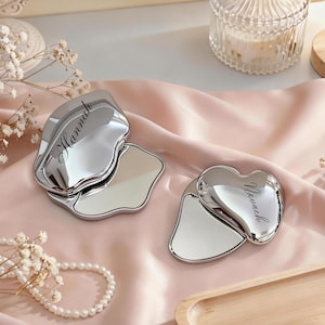 May include: Two silver-colored gua sha tools on a pink satin surface. One is a wavy shape with "Hannah" engraved, the other is heart-shaped with "Neveah" engraved. The tools are designed for facial massage and skincare.