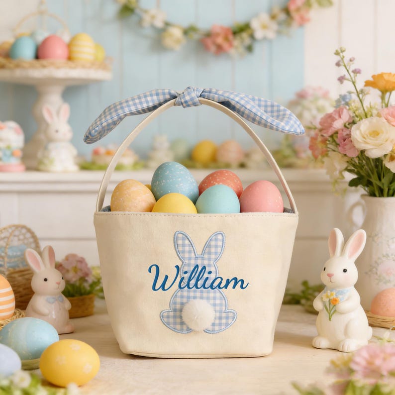 Pu&ograve; includere: Un cestino pasquale color crema con manico e fiocco a quadretti blu, pieno di uova dai colori pastello. Il cestino presenta un'applicazione di coniglietto blu e il nome "William". Coniglietti e uova in ceramica decorano lo sfondo.