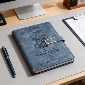 K&ouml;nnte beinhalten: Blaues Lederjournal mit goldenem Verschluss. Auf dem Einband steht der Text "THE NEXT Chapter" &uuml;ber einem floralen Design und dem Namen "SARAH WILSON". Auf dem unteren Teil des Journals steht "Each chapter ushers in new growth". Ein blauer Stift liegt auf dem Schreibtisch.