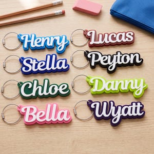 Può includere: Otto portachiavi con nomi personalizzati dotati di anelli in metallo argentato, ognuno con un nome in corsivo bianco in rilievo su sfondo colorato. I nomi sono Henry, Lucas, Stella, Peyton, Chloe, Danny, Bella e Wyatt in blu, bordeaux, blu scuro, nero, verde, verde lime, rosa e viola.