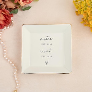 May include: A square, off-white trinket dish with a gold rim. The dish has the words "sister EST. 1995 aunt EST. 2025" and a heart symbol printed in black. The dish is on a light pink surface.