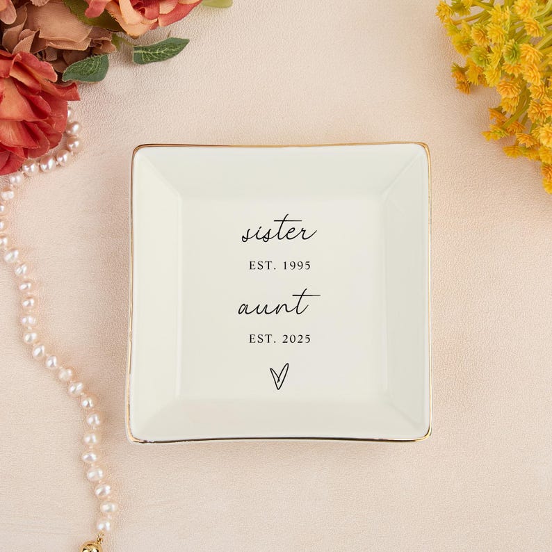 May include: A square, off-white trinket dish with a gold rim. The dish has the words "sister EST. 1995 aunt EST. 2025" and a heart symbol printed in black. The dish is on a light pink surface.