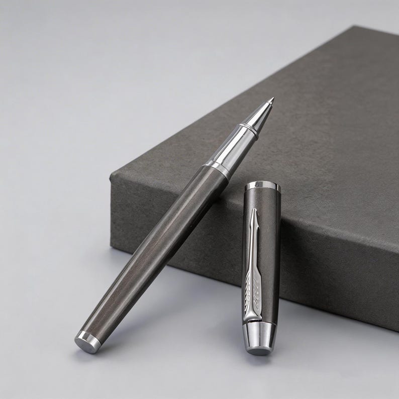 May include: A dark gray rollerball pen with silver accents, cap removed and placed next to the pen. The pen is resting on a dark gray box. The pen's tip and cap are silver.