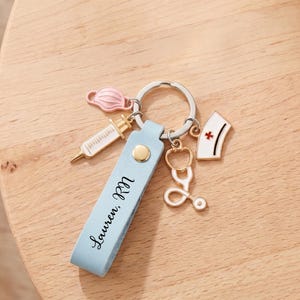 Pu&ograve; includere: Un portachiavi in pelle azzurra con il nome "Lauren, RN" stampato sopra. Il portachiavi ha un anello argentato e include ciondoli di una siringa, una mascherina, uno stetoscopio e un cappello da infermiera.