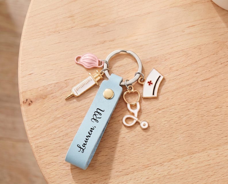 Pu&ograve; includere: Un portachiavi in pelle azzurra con il nome "Lauren, RN" stampato sopra. Il portachiavi ha un anello argentato e include ciondoli di una siringa, una mascherina, uno stetoscopio e un cappello da infermiera.