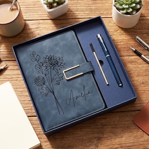 May include: A blue notebook with a black floral line art design and the name Amelia printed on the cover, featuring a gold-toned metal clasp. The notebook is presented in a matching blue gift box alongside a blue ballpoint pen and a metal refill.