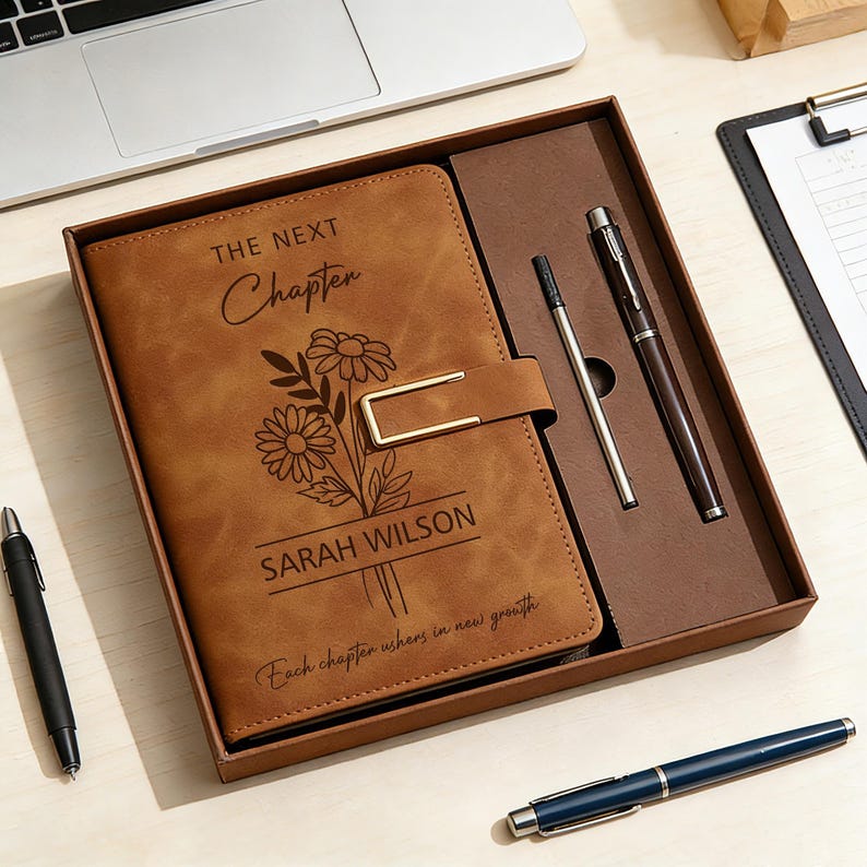K&ouml;nnte beinhalten: Ein braunes Lederjournal mit den Worten "The Next Chapter" und "Sarah Wilson" auf dem Einband sowie einem floralen Design. Das Journal befindet sich in einer Geschenkbox mit zwei Stiften. Ein schwarzer Stift und ein blauer Stift sind ebenfalls sichtbar.