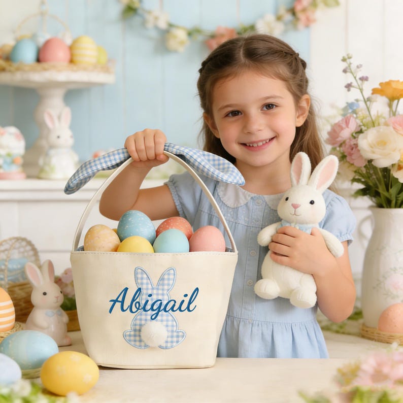 Pu&ograve; includere: Un cestino pasquale beige con un design a coniglietto a quadretti blu e il nome "Abigail" in blu. Il cestino &egrave; pieno di uova dai colori pastello. Un coniglietto di peluche bianco &egrave; tenuto da una persona. Altre decorazioni pasquali sono sullo sfondo.