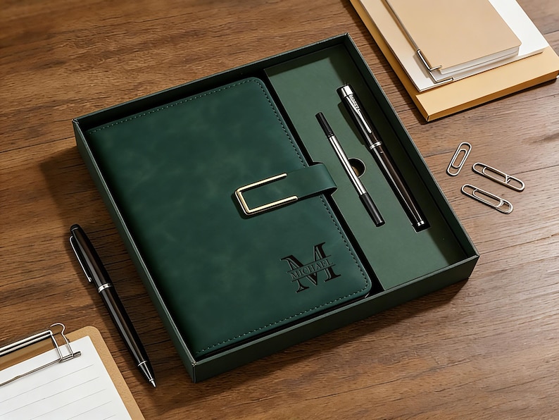 Op de afbeelding: Een donkergroene notitieboekenset met een gouden sluiting, twee pennen en paperclips in een bijpassende doos. Het notitieboek is voorzien van de naam "MICHAEL XVI". De set wordt gepresenteerd op een houten oppervlak.