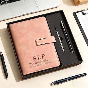 May include: A pink journal with a gold clasp, personalized with "SLP" and a name, comes with two pens in a black box. A black pen and a blue pen are also visible.