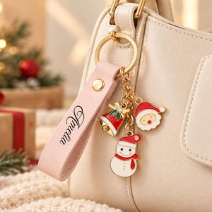 May include: A light pink leather keychain strap personalised with the name Amelia in black script, attached to a gold metal ring featuring three dangling charms: a red and white bell, a Santa Claus face, and a snowman in a red hat.