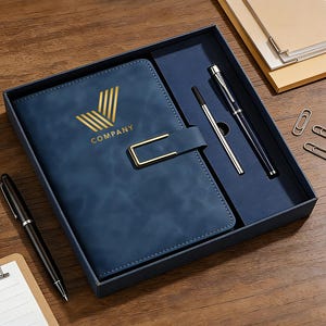 Puede incluir: Un cuaderno azul con cierre dorado y la palabra "COMPANY" impresa en la portada. El cuaderno está en una caja de regalo con dos bolígrafos y clips. El conjunto está sobre una superficie de madera.