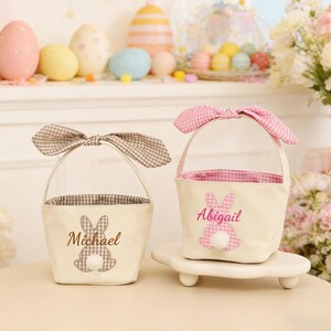 Pu&ograve; includere: Due cestini pasquali color crema con manici a forma di orecchie di coniglio. Un cestino ha un fiocco a quadretti marrone e il nome "Michael". L'altro ha un fiocco a quadretti rosa e il nome "Abigail". Entrambi hanno dettagli di coda di coniglio.