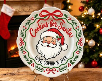 May include: A white ceramic plate with a scalloped edge featuring a central illustration of Santa Claus. The text reads Cookies for Santa and Love, Sophia & Jack. The design includes a green holly wreath border with red bows and berries.
