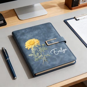 May include: A blue journal with a yellow flower illustration and the name "Evelyn" in white script. The journal has a gold-colored clasp and a matching blue pen is next to it. The journal is on a gray desk.
