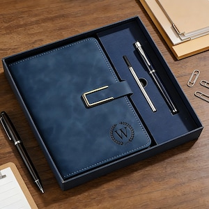 Op de afbeelding: Een blauwe notitieboekenset van imitatieleer met een gouden sluiting, een pen en een zilveren pen. Het notitieboek heeft een monogram "W" in een lauwerkrans. De set zit in een donkerblauwe doos, met paperclips en een notitieblok.
