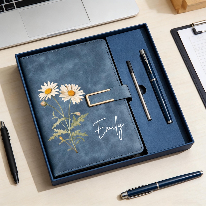 May include: A blue leather journal with a daisy floral design and the name "Emily" in white script. The journal is in a gift box with two pens. The journal has a gold-colored clasp.