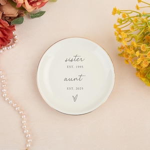 May include: A cream ceramic trinket dish with a gold edge. The dish is inscribed with the words "sister" and "aunt" in cursive, along with the years "EST. 1995" and "EST. 2025", and a heart symbol.