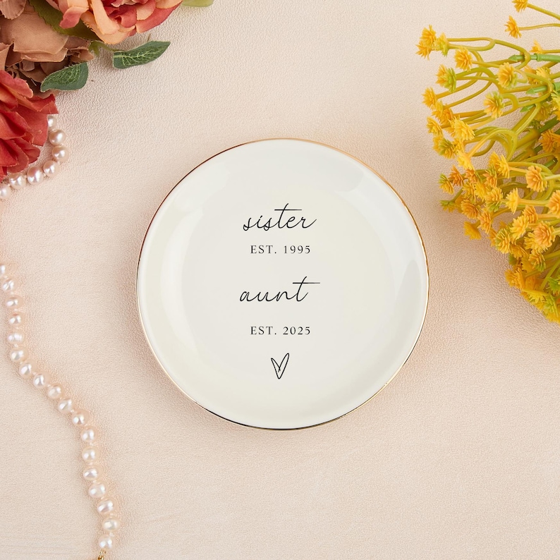 May include: A cream ceramic trinket dish with a gold edge. The dish is inscribed with the words "sister" and "aunt" in cursive, along with the years "EST. 1995" and "EST. 2025", and a heart symbol.