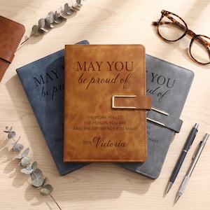 Könnte beinhalten: Ein braunes Notizbuch in Lederoptik mit einem goldfarbenen Metallverschluss, geprägt mit dem Text: MAY YOU be proud of THE WORK YOU DO THE PERSON YOU ARE AND THE DIFFERENCE YOU MAKE FOR Victoria. Zwei weitere Notizbücher in Blau und Grau sind sichtbar.