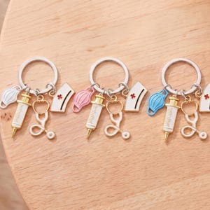Pu&ograve; includere: Tre portachiavi con ciondoli a tema medico. Ogni portachiavi ha un anello argentato e presenta una siringa, uno stetoscopio, un cappello da infermiera e una mascherina. Le mascherine sono bianche, rosa e blu. I cappelli da infermiera hanno una croce rossa.