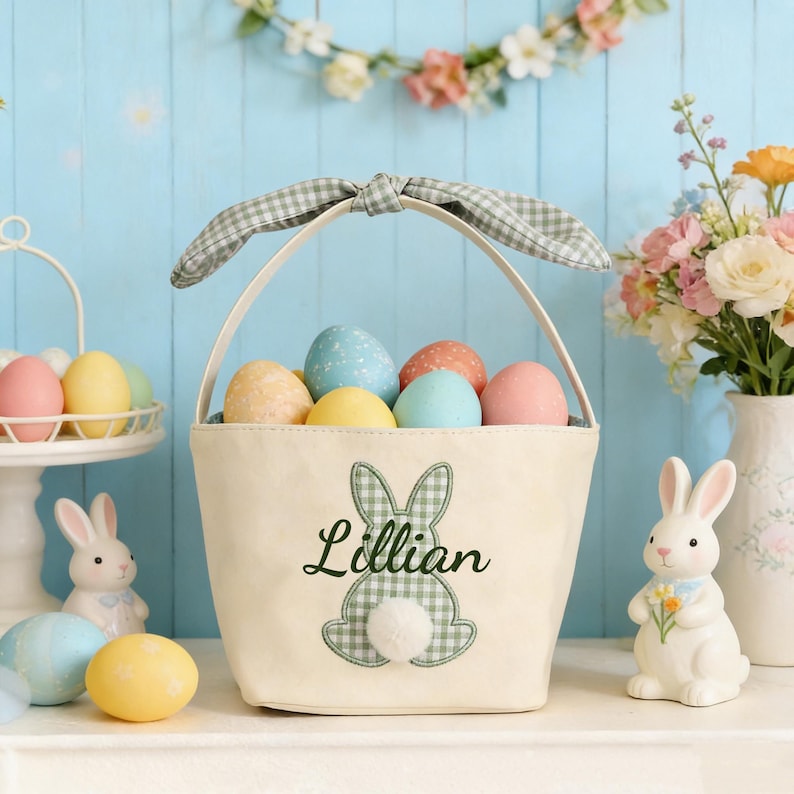 Pu&ograve; includere: Un cestino pasquale color crema con manico a fiocco a quadretti, pieno di uova dai colori pastello. Il cestino presenta un design a coniglietto a quadretti verdi e il nome "Lillian". Sono presenti anche coniglietti in ceramica e una composizione floreale.