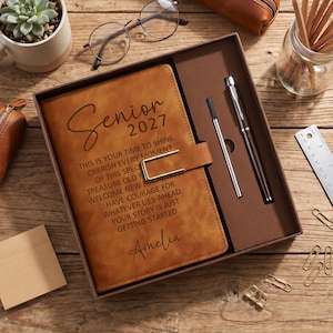 May include: A tan leather-like journal in a gift box with a metal clasp, featuring the text Senior 2027, an inspirational quote, and the name Amelia. The set includes a silver pen and a thin metal stylus, presented in a brown tray.