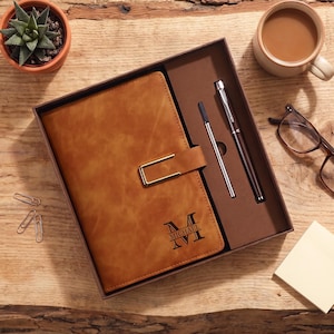 Op de afbeelding: Een bruin notitieboek in lederlook met een goudkleurige metalen sluiting en de naam MICHAEL gedrukt over een grote letter M. Het notitieboek zit in een bruine geschenkdoos samen met een zilverkleurige pen en een dunne metalen vulling.