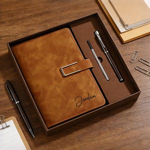 Op de afbeelding: Een bruin leren notitieboek met een metalen sluiting, de naam "Jordan" staat op de voorkant. Het notitieboek zit in een geschenkdoos met twee pennen, paperclips en een notitieblok. Het notitieboek en de pennen zijn bruin.