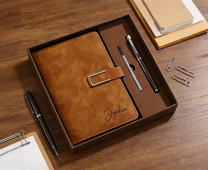 Op de afbeelding: Een bruin leren notitieboek met een metalen sluiting, de naam "Jordan" staat op de voorkant. Het notitieboek zit in een geschenkdoos met twee pennen, paperclips en een notitieblok. Het notitieboek en de pennen zijn bruin.