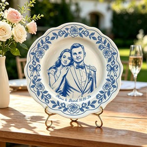 May include: A white ceramic decorative plate with a scalloped edge featuring a blue ink-style illustration of a couple. The border displays roses and bows. The text "Olivia & David 10.05.26" is printed below the couple. The plate rests on a gold stand.