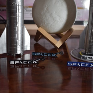 May include: Two silver model rockets with black and white SpaceX logos on their bases. The rockets are on a wooden table with a moon lamp in the background.