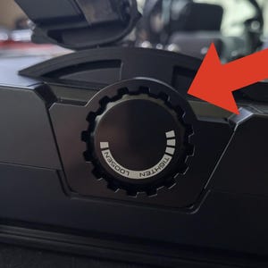 May include: A black circular adjustment knob on a mechanical device featuring a gear-like edge. The knob includes white printed text that reads TIGHTEN and LOOSEN, along with a series of white rectangular indicators to show the rotation direction.