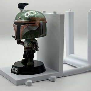 May include: A white plastic display stand with two vertical supports and a base. A Star Wars Boba Fett Pop figure is displayed on the base. The figure is green, brown, and black with a helmet and a blaster.
