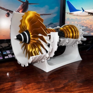 May include: A white and gold model of a jet engine with a fan blade and a stand. The engine is on a wooden surface.