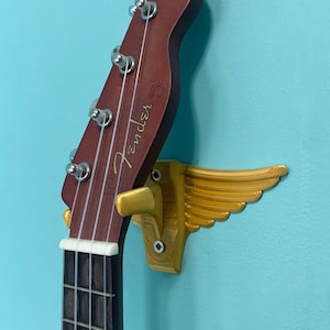 May include: A brown ukulele with the Fender logo, mounted on a gold-colored wall hanger shaped like wings. The ukulele has silver tuning pegs and a dark fretboard. The wall is painted a light blue.