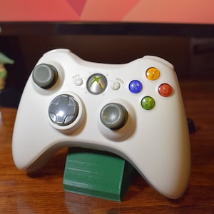 May include: A white Xbox 360 controller with green, blue, orange, and yellow buttons. The controller is sitting on a green stand.