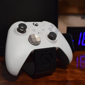 May include: A white Xbox controller with a black stand. The controller has a black faceplate with a gray button. The stand has the Xbox logo on it.