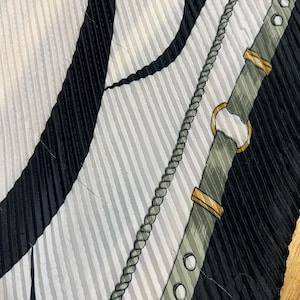 May include: A close-up of a pleated silk scarf with a pattern of black and white stripes, a gray rope design, and a gray belt with gold-colored accents. The scarf has a textured, ribbed appearance.