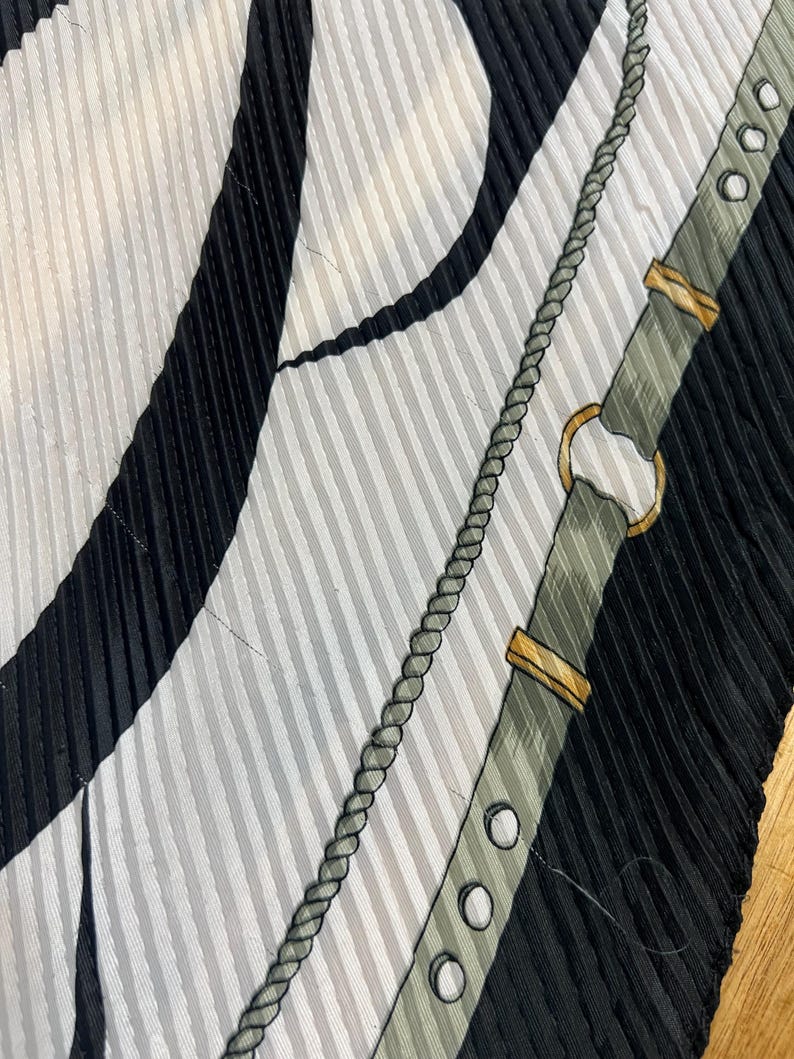 May include: A close-up of a pleated silk scarf with a pattern of black and white stripes, a gray rope design, and a gray belt with gold-colored accents. The scarf has a textured, ribbed appearance.